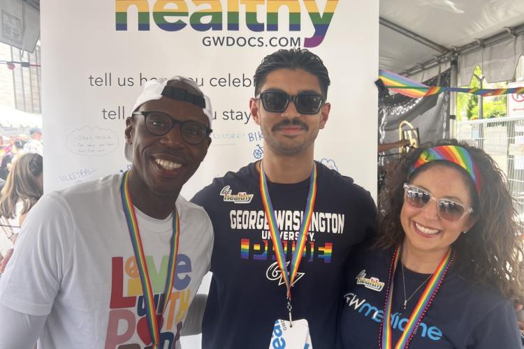 A black man, a brown man, and a black woman stand together in workout clothes, smiling, in front of sign that says BE HEALTHY
