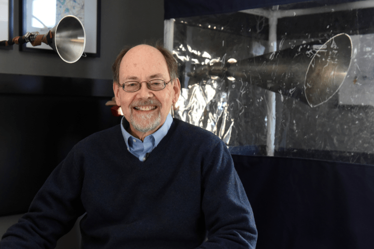 Professor in glasses, wearing blue v-neck and light blue shirt, sits in front of the gesundheit machine, which he invented