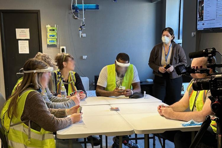 Volunteer flu study participants sit around a table and play a game, some are wearing protective face shields, some are not