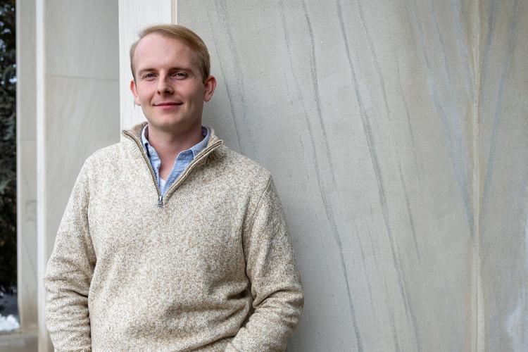 White man with blond hair in beige top standing in front of marble wall looks to camera with a small smile