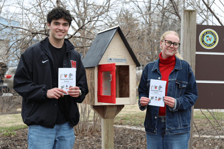 Two students hold recipe book they created, standing in front of a wooden library at Hyattsville Food Forest