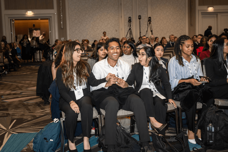 Three students sit smiling, holding hands in a conference room full of people