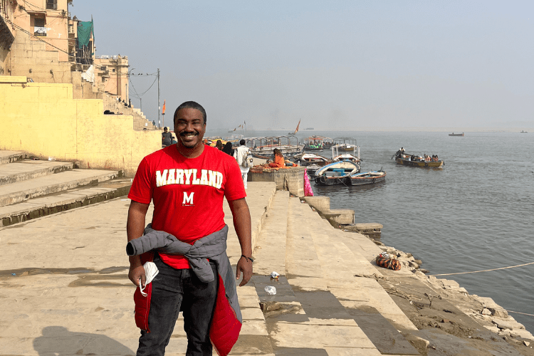 Black man in red MARYLAND shirt and black trousers stands smiling beside the Ganges river in India