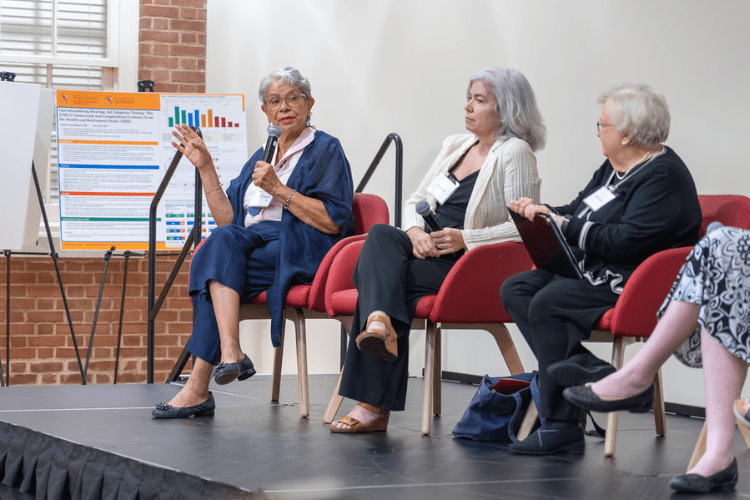 Three elder women sit on stage red chairs with a poster behind them displaying age health research