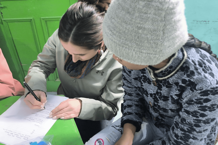 young indian pupil watches as UMD student writes on her paper in a classroom in Varanasi, the table is painted bright green and the walls are light blue