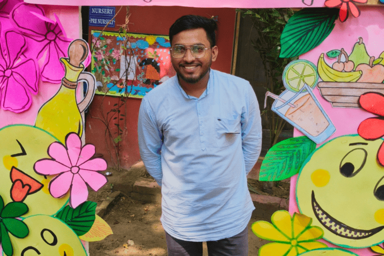 Indian man smiles with his hands behind his back, standing in between large paper flowers and smiley faces