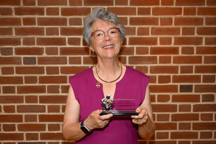 An older woman in glasses short grey hair and purple top smiles to camera holding an award