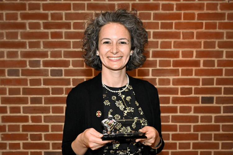 Woman with greying curly hair pulled back smiles to camera holding an award