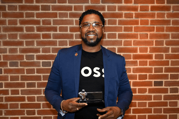 Black man in blue suit and black tshirt with short hair and sunglasses smiles to camera holding an award