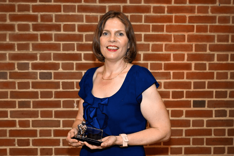 White woman in blue dress holds award, looking at camera