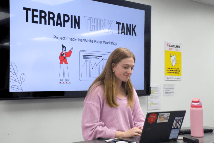 Student with blond hair works at laptop in front of big screen with words TERRAPIN THINK TANK
