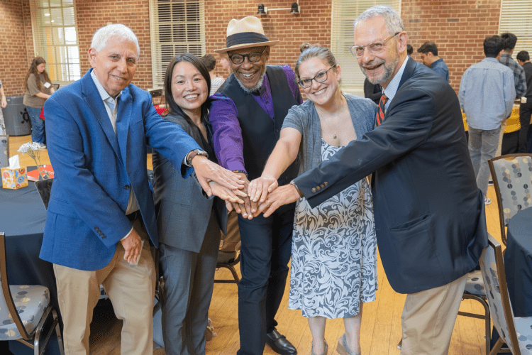 group of publihealth researchers pose with one arm reached out in front to touch hands, smiling, in a brick walled conference room