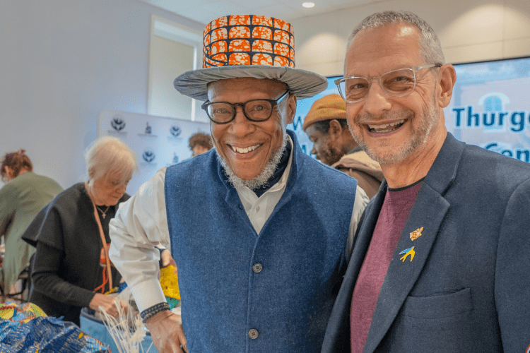 Black male professor wearing top hat made from African fabric poses with white male Dean in a blue sports jacket also smiling