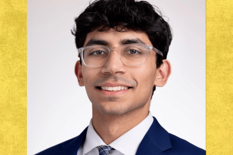 Student wearing dark blue suit jacket, white shirt and colour tie, brown wavy short hair, with glasses on smiles to the camera