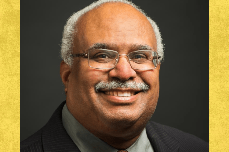 Black older man with greying moustache and rimless spectacle smiles to camera, wearing a dark suit jacket and reddish tie