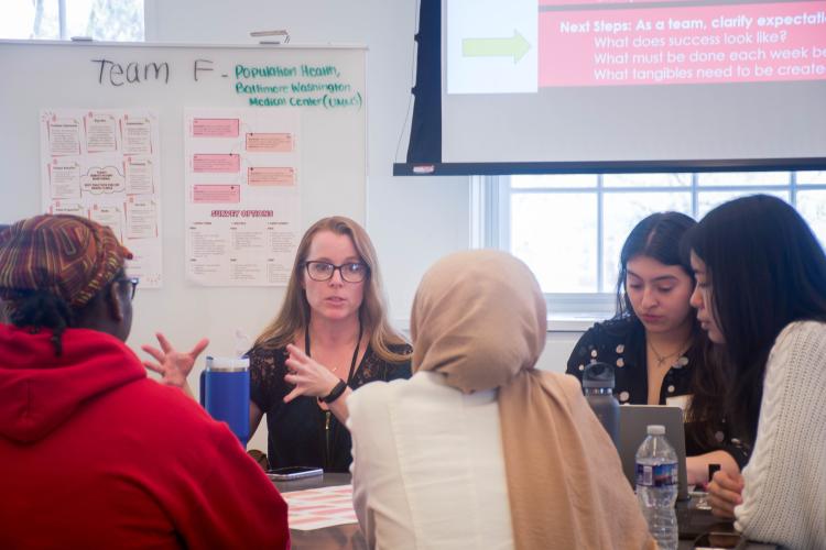 Professional woman with black glasses speaks to group of students with her hands raised in a gesture, we see 3 students from behind, one student’s face and in background a white board with words on it