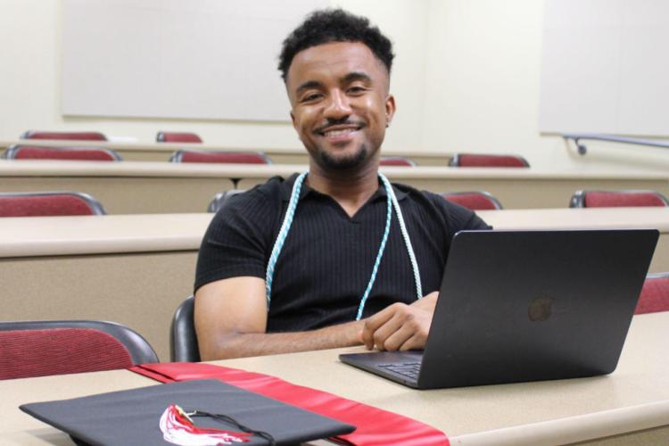 Black male grad student sitting in classroom with laptop and graduation sash, smiles to camera