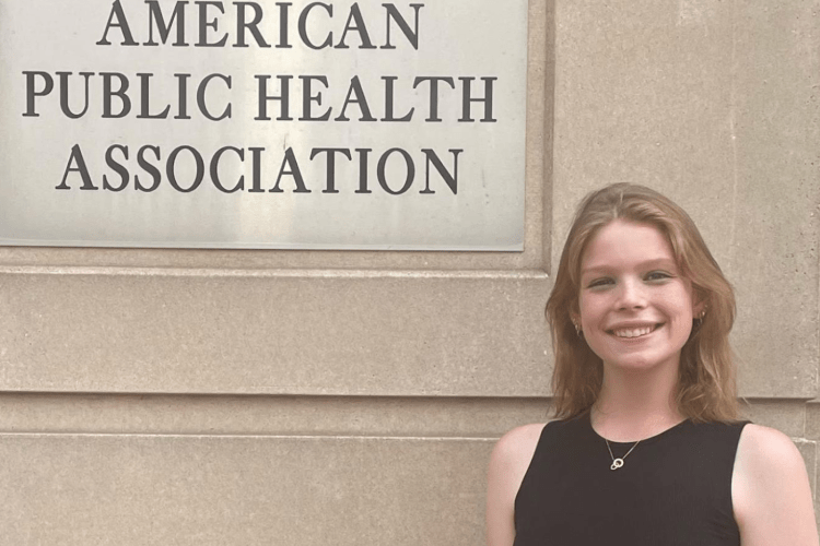 Young white woman in black tank top smiles to camera next to sign for the American Public Health Association