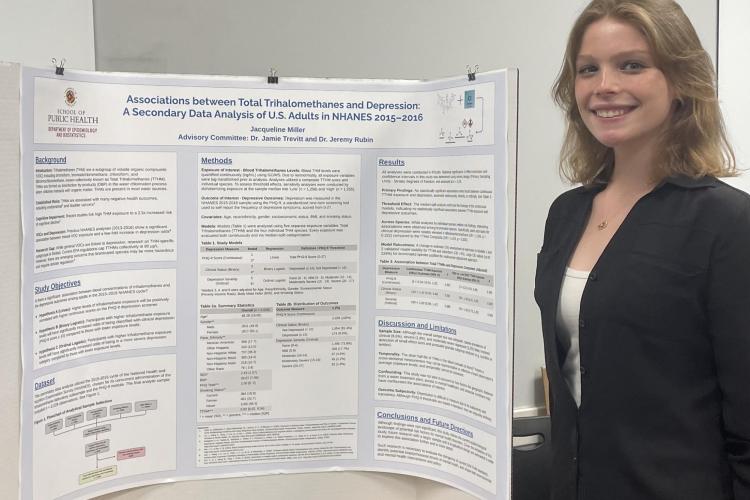 young woman in a dark suit and white top stands smiling next to a public health presentation board