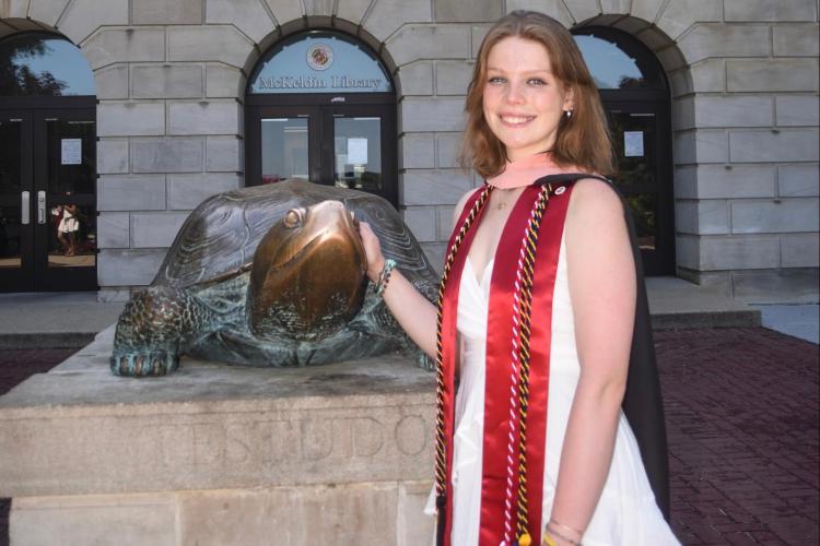 young woman wearing white dress and red graduation scarf touches the nose of a bronze turtle statue called “Testudo"