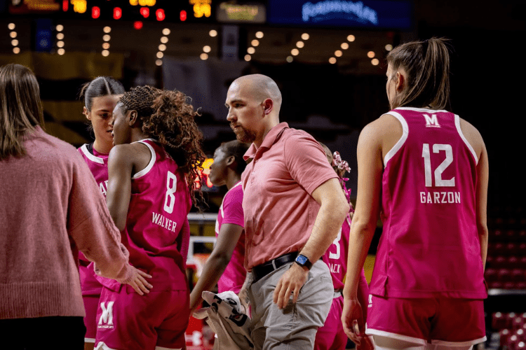 Man in pink shirt and khaki slacks walks between women basketball players court side
