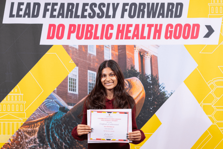 Wearing a dark red top, student with long brown hair smiles to camera, standing in front of SPH banner and holding her award certificate