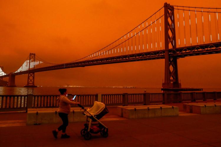 red smokey sky behind a big bridge, in foreground a person pushes a stroller while looking at their phone