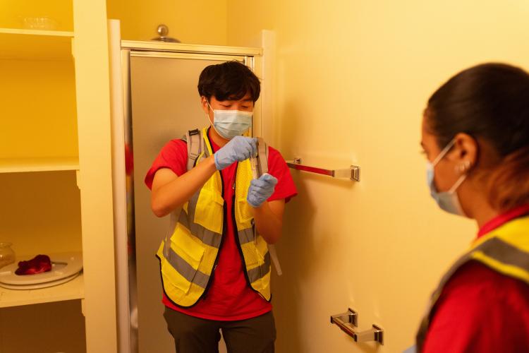 Young man in hazard vest and facemask takes water sample in the basement of a house, with another student in face mask and vest watching