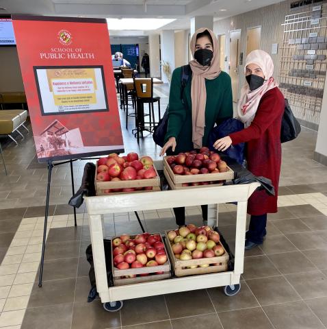Two students select fresh fruit from a cart filled with apples.