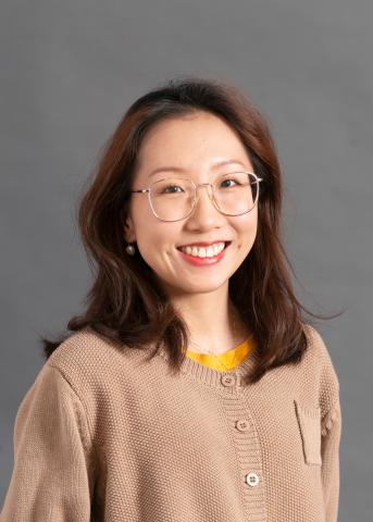 Asian woman in beige cardigan and glasses smiles at the camera, with grey background
