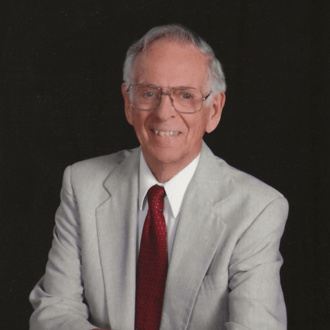 Portrait of Dr. David Clarke seated in a light gray suit and red tie against a dark background.