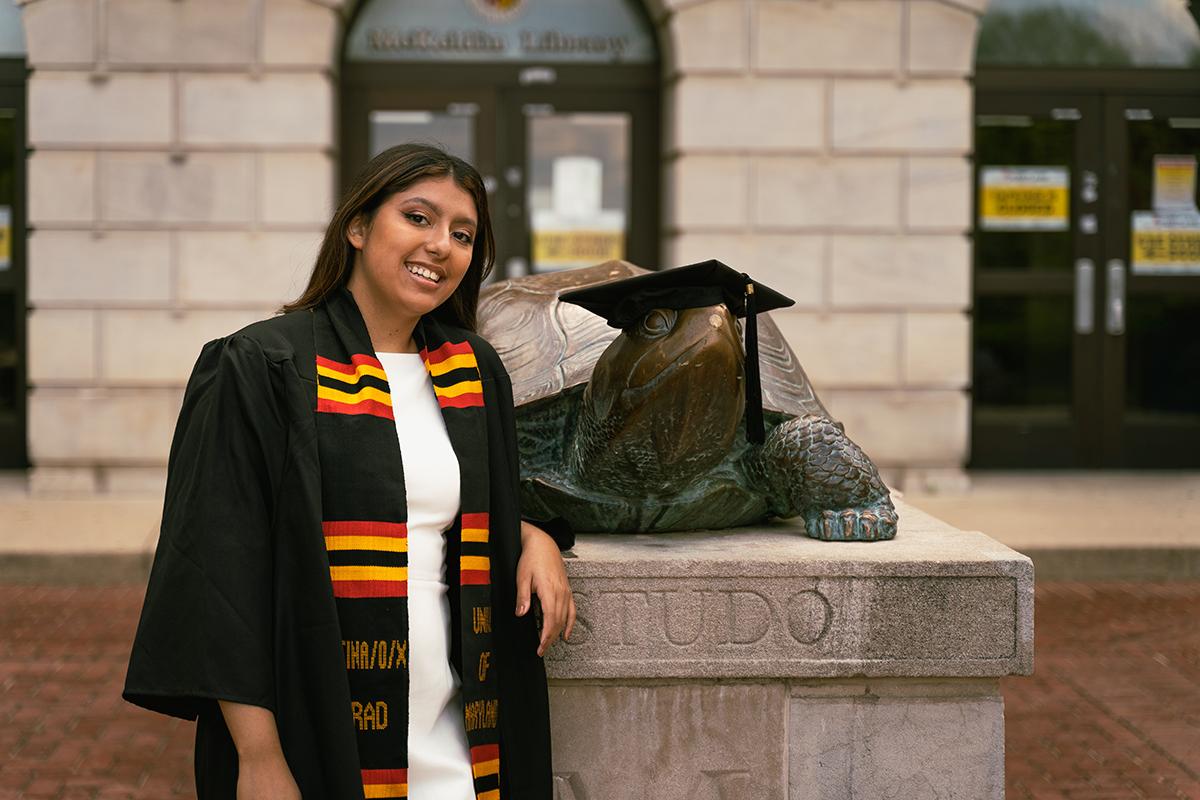 Stephanie Bautista, a UMD School of Public health graduate leans on the Testudo statue outside of McKeldin mall with her commencement regalia on