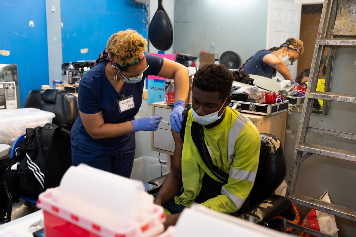 Black female nurse standing up and giving COVID vaccine in the arm of a young Black man who is seated and wearing construction worker clothing.