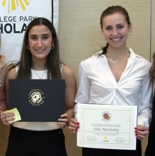 Two female students stand holding award certificates