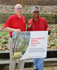 A man and woman wearing red shirts stand behind a sign announcing a new heart discovery trail.