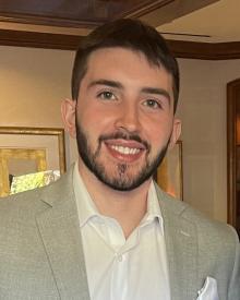 Angelo is smiling at the camera. He has short hair and a short beard with mustache.