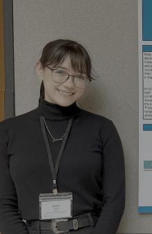 Jenna has her hair pulled into a pony tail with bangs, she is smiling at the camera wearing glasses.