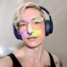 Headshot of Nora Jameson. Nora is wearing headphones and a rainbow light reflected in her face.
