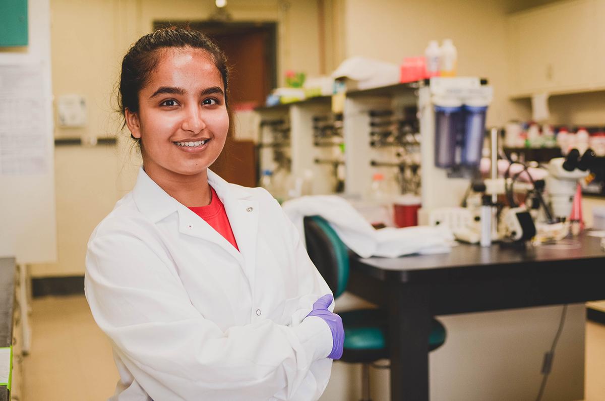 Female student in white lab coat smiling with laboratory in background.