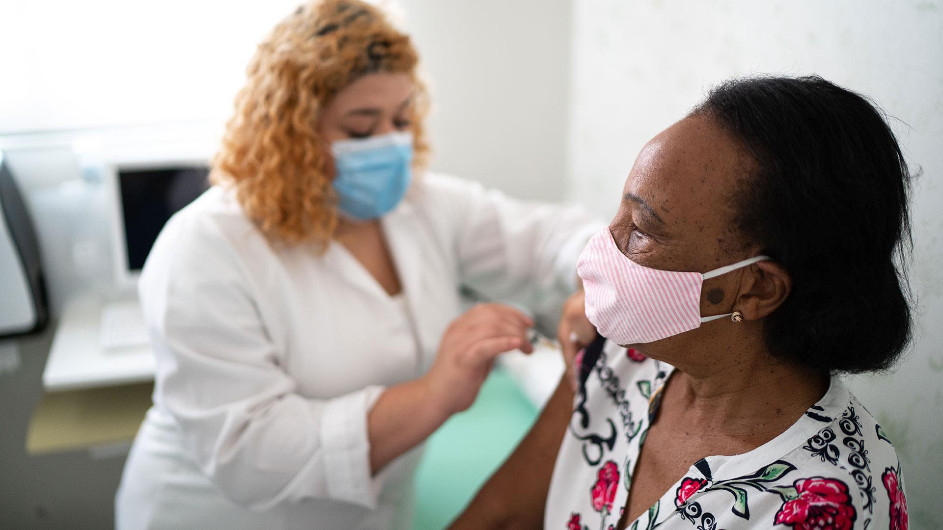 African American older woman getting vaccinated by another woman, both wearing masks to protect from COVID-19