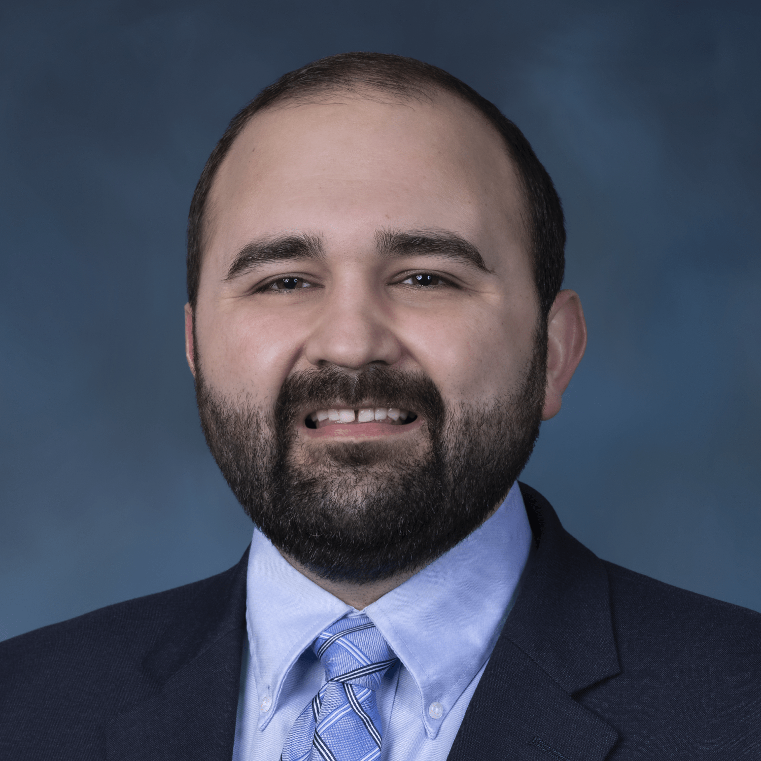 Close up of Martin Gesheff with black mustache and beard and wearing a black suit, baby blue shirt and a patterned tie