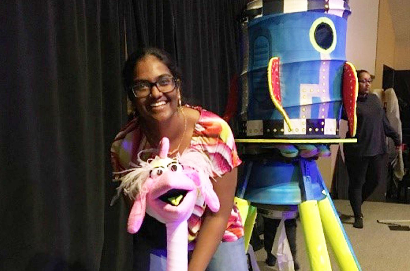 Student from the University of Maryland wearing glasses and smiling at camera poses with llama puppet