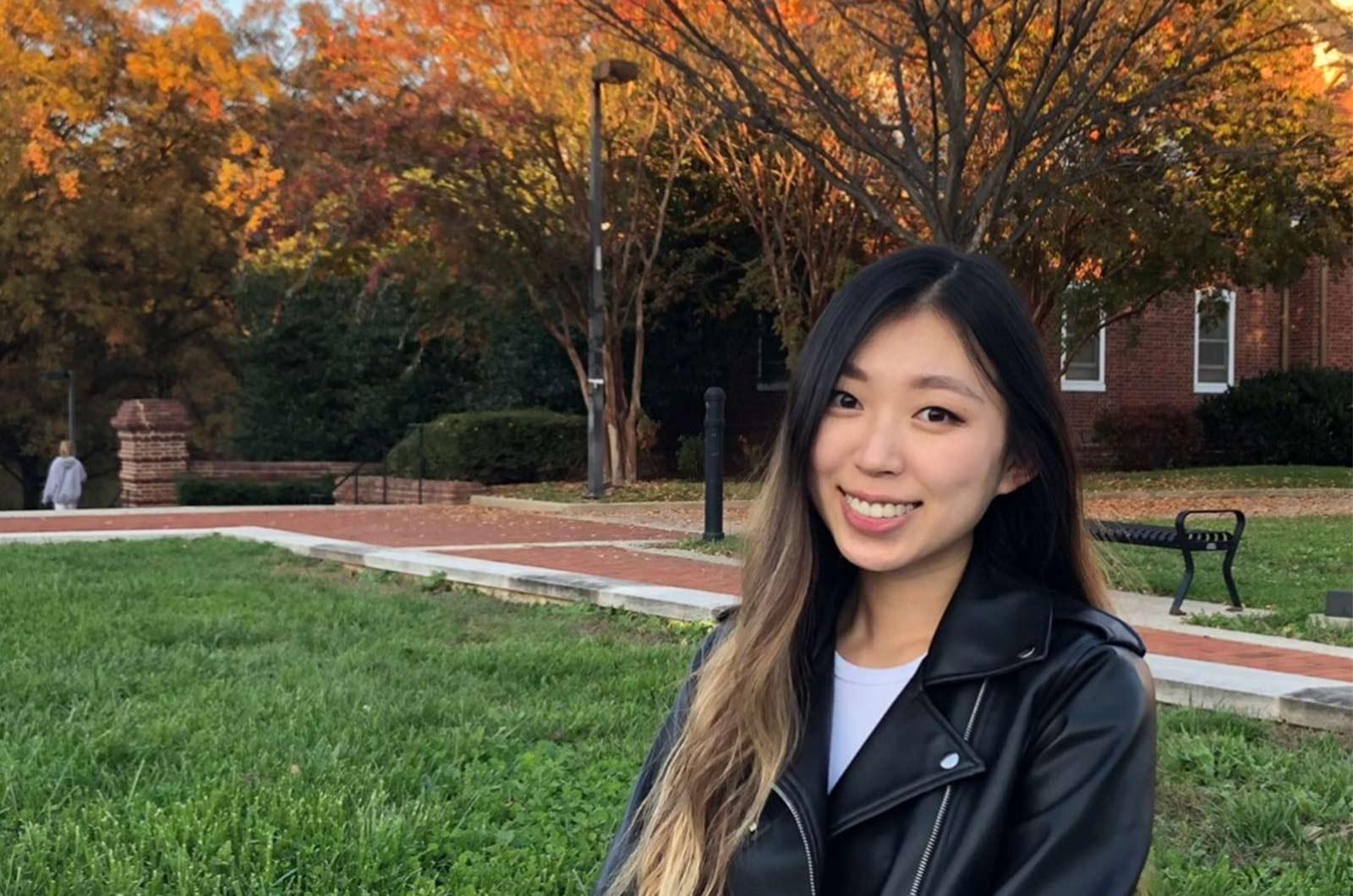 Student from the University of Maryland smiles at camera, posed in front of UMD building, trees with fall foliage