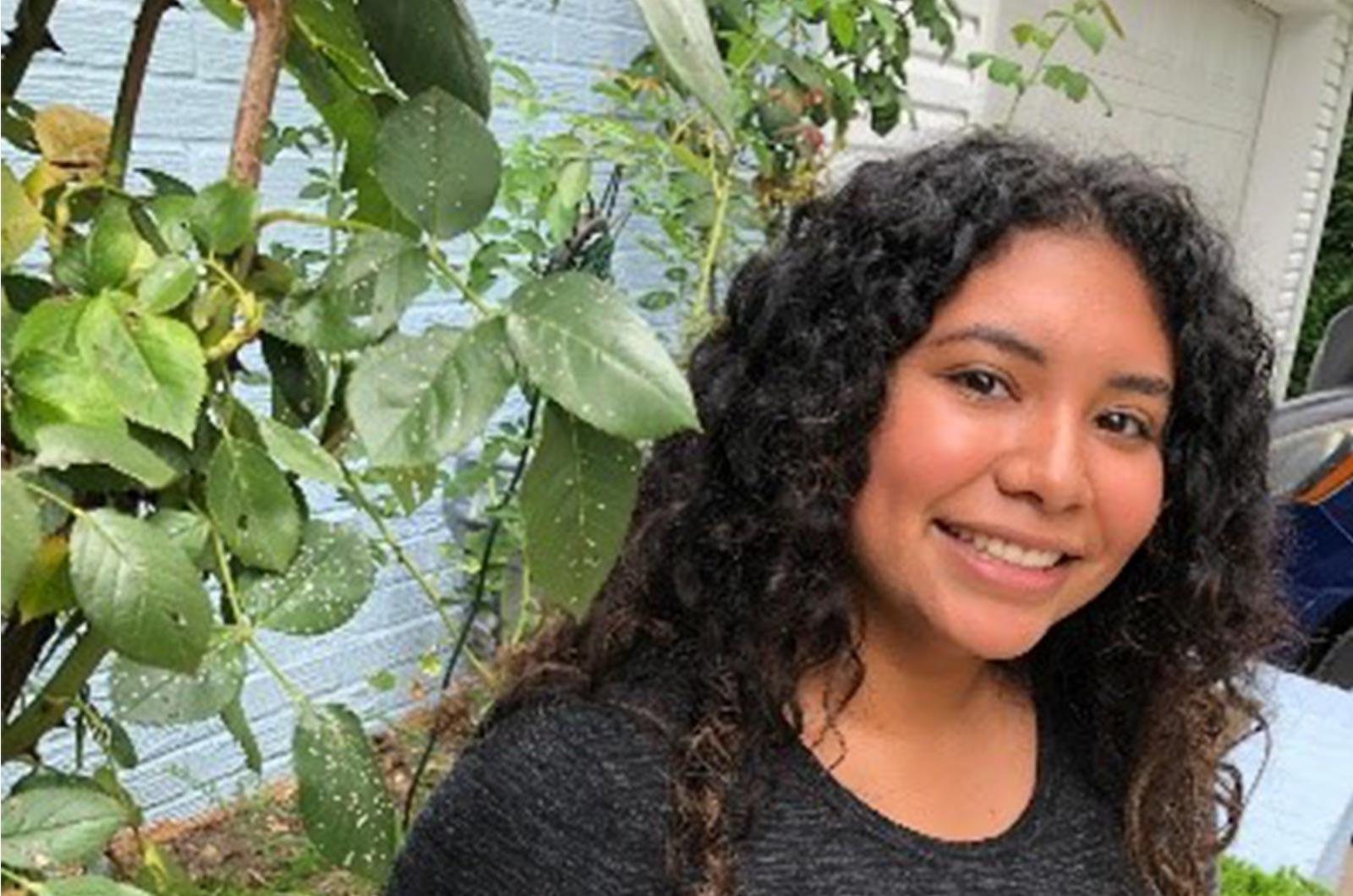 Student from the University of Maryland smiles at camera in front of plants and painted brick wall