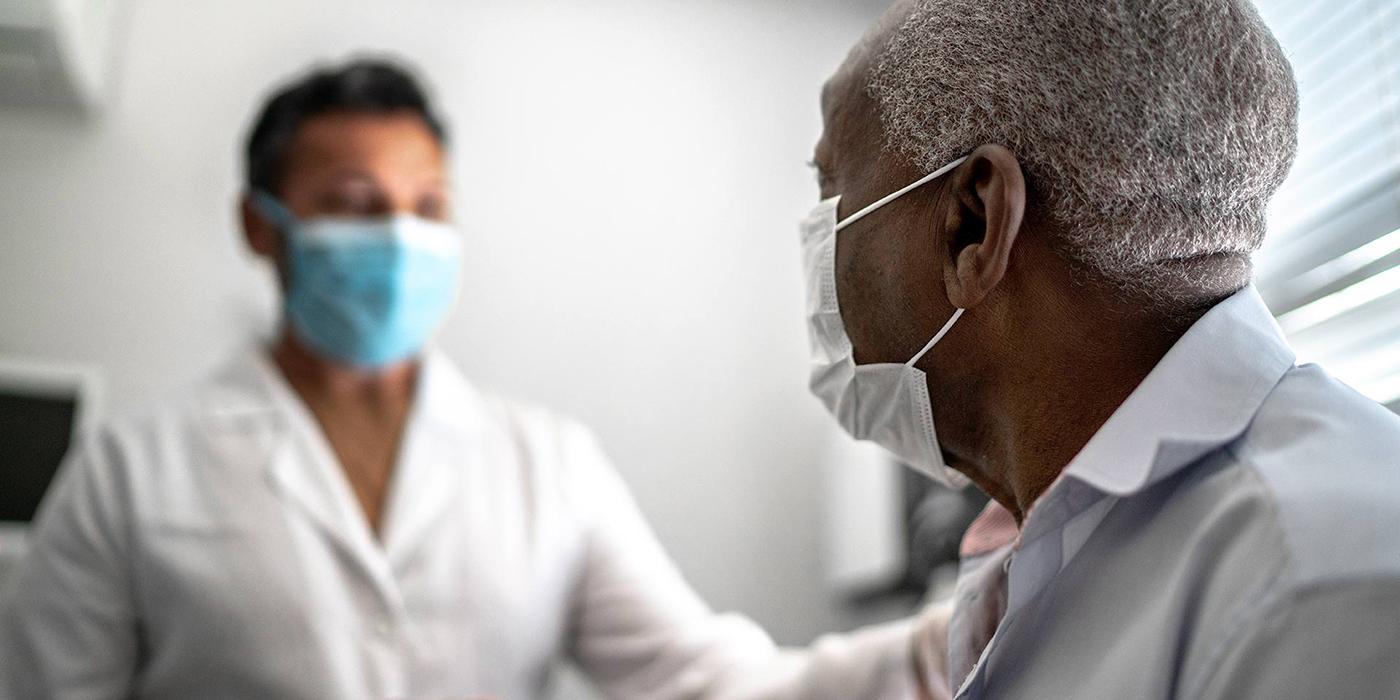 Older African American man seated in doctor's office with health care professional standing up, both are wearing masks.