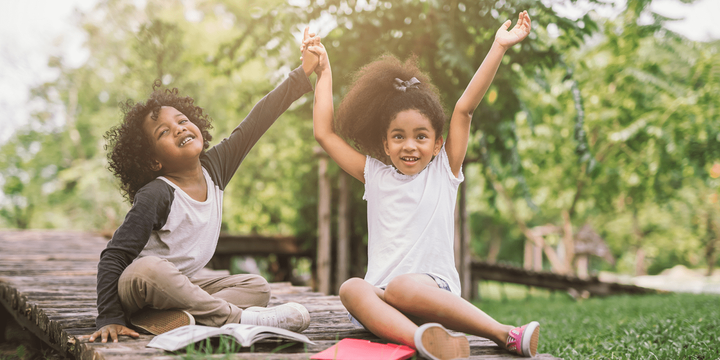 Two Black children sit outside during summer looking happy with arms in the air