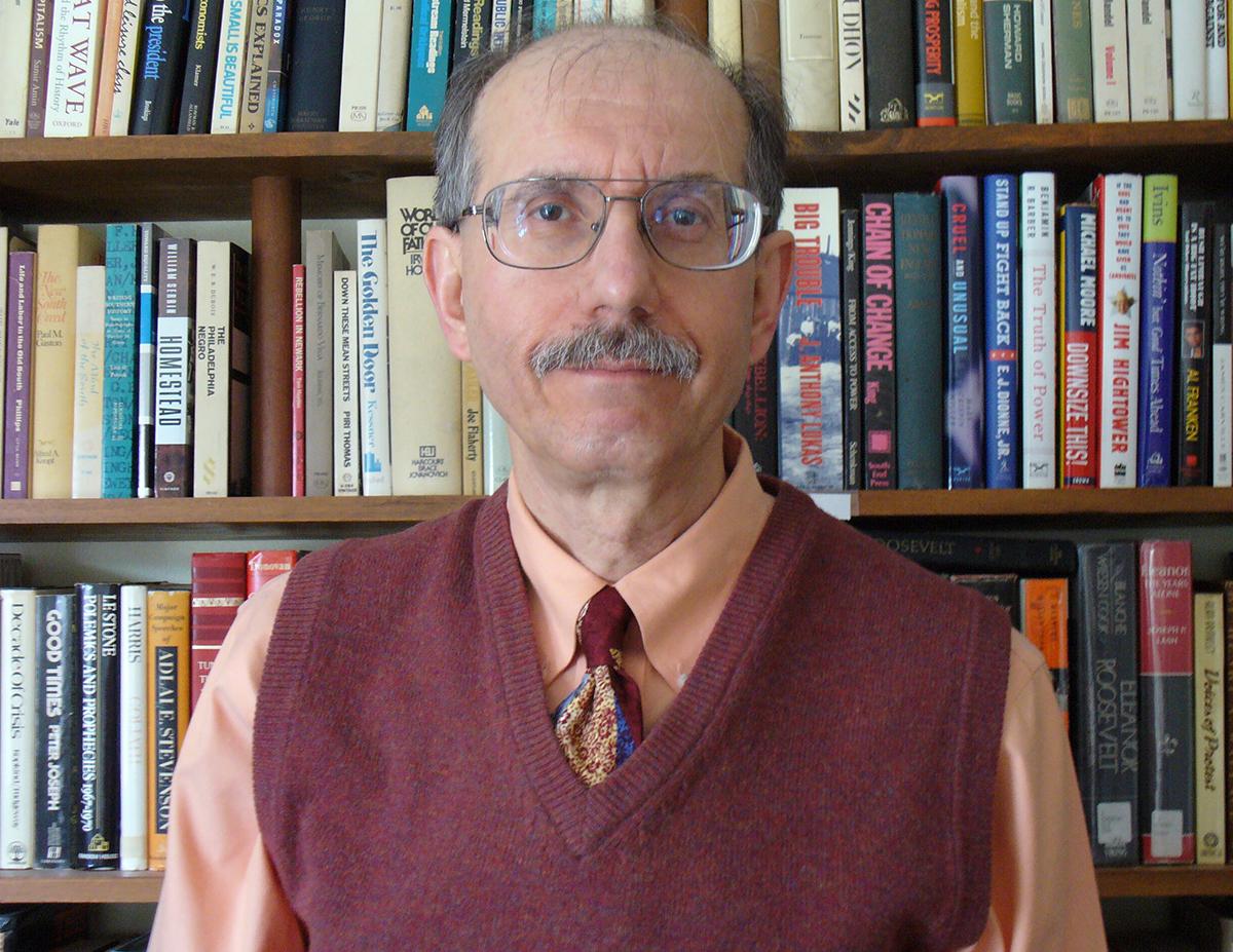 Sam Pizzigati, an older white man with a mustache, bald head and glasses wearing a sweater vest, shirt and tie in front of a bookshelf
