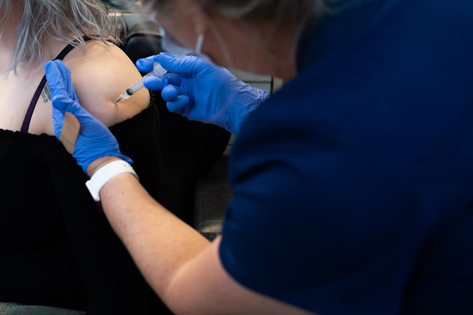 Close up of White woman getting vaccinated by a woman wearing blue gloves. Shoulder tattoo with a crown and a date is visible.