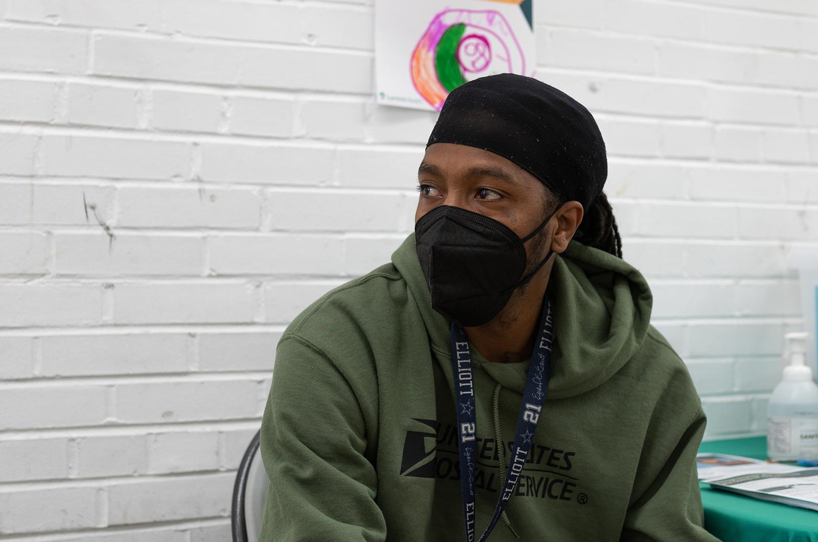 Close up of a Black man looking off to the side and wearing a US Postal Service sweatshirt, a do rag, and a black facial barrier (for COVID-19) 
