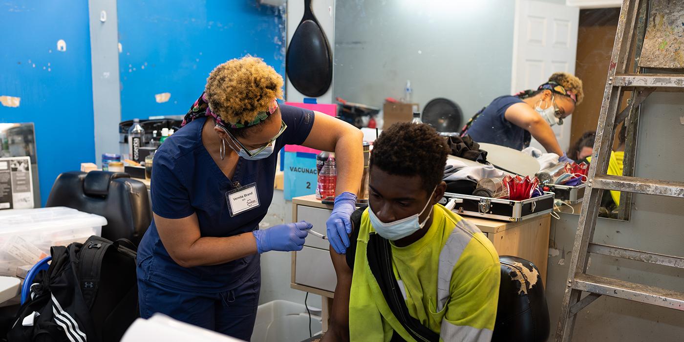 Black female nurse standing up and giving COVID vaccine in the arm of a young Black man who is seated and wearing construction worker clothing.